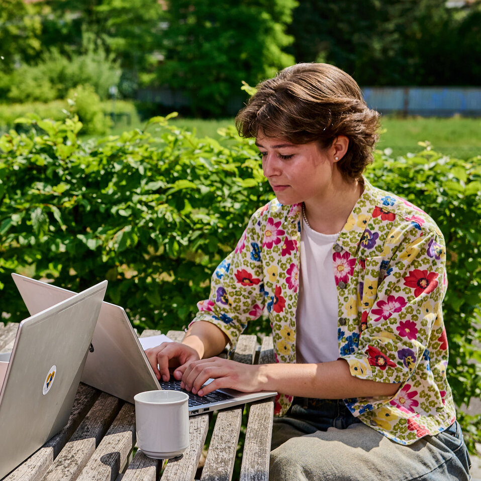 Zwei Studierende sitzen am Tisch im Innenhof der KHSB und arbeiten an ihren Laptops.