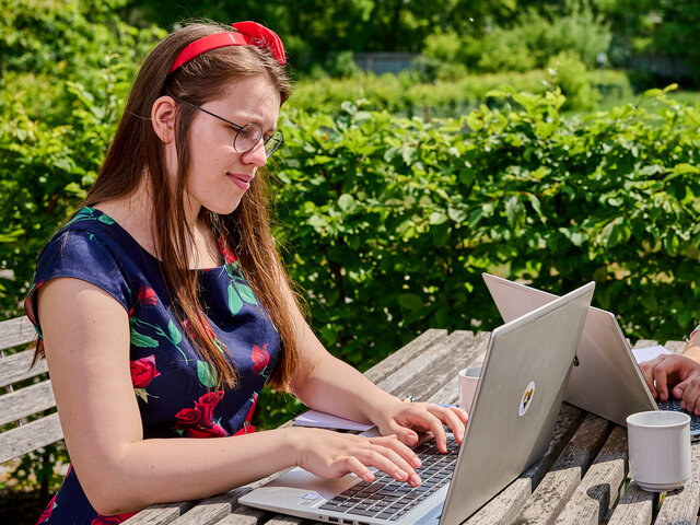 240527_KHSB_Karriere_Boillot_48 Zwei Studierende sitzen am Tisch im Innenhof der KHSB und arbeiten an ihren Laptops.
