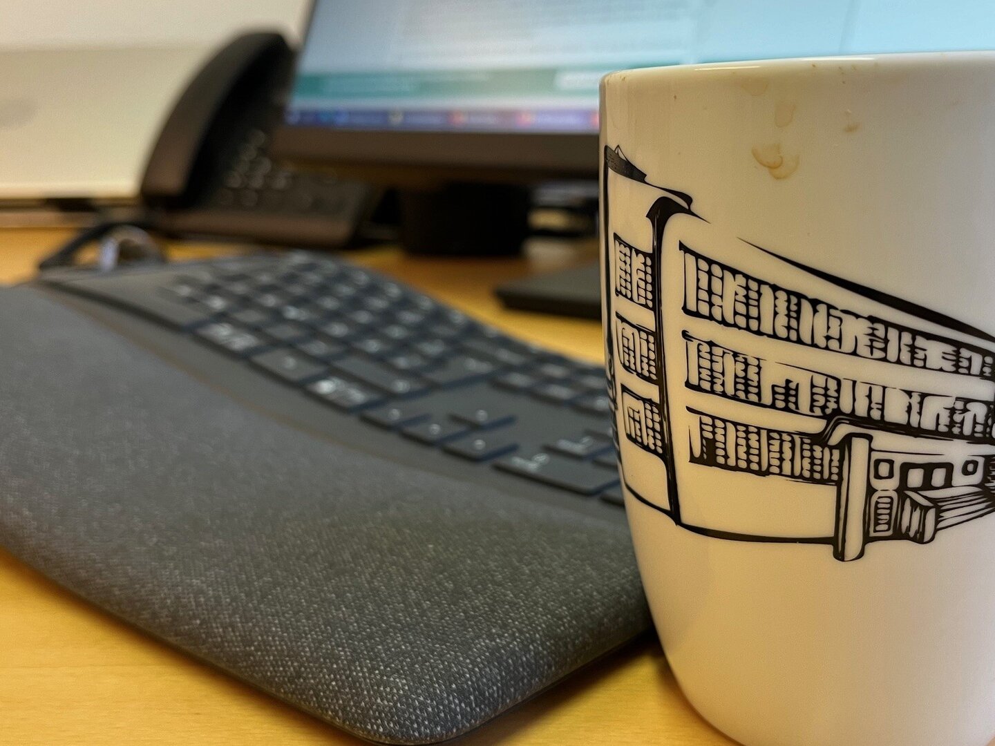 Foto mit Nahaufnahme eines Schreibtisches, im Vordergrund ist eine Tasse mit KHSB Werbeaufdruck, im Hintergrund sind Tastatur, Bildschirm und Laptop zu sehen.