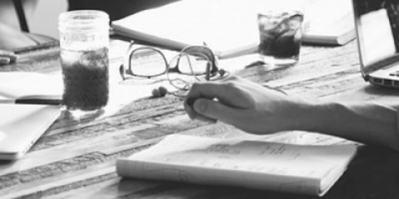 Mehrere Personen mit aufgeschlagenen Notizbüchern und Laptop am Schreibtisch im Gespräch