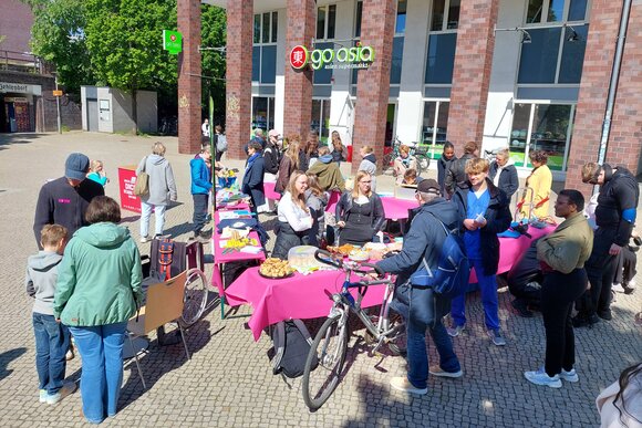 Tische mit pinker Tischdecke im Sonnenschein auf einem großen Platz. Man sieht zahlreiche Menschen, die sich an den Tischen zum Thema Pflege informieren. Auf einem Tisch steht Kuchen.