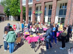 Tische mit pinker Tischdecke im Sonnenschein auf einem großen Platz. Man sieht zahlreiche Menschen, die sich an den Tischen zum Thema Pflege informieren. Auf einem Tisch steht Kuchen.