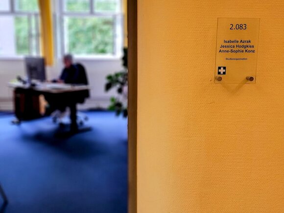 Ersthelferkreuz auf Türschild an der KHSB, im Hintergrund sitzt Person am Schreibtisch