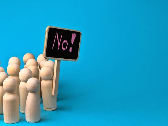 Im Bild wird ein Protest mit Holzfiguren in einer Menge und einem Schild mit der Aufschrift 'No!' dargestellt