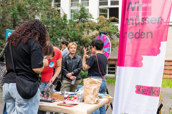 Menschen unterhalten sich an einem mit Bastelmaterialien vollgestellten Tisch unter freiem Himmel. Im Hintergrund ein Glücksrad. Im Vordergrund eine Fahne mit der Aufschrift "Wir müssen reden".