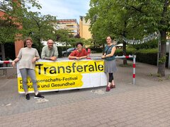 Ein gelbes Banner mit der Aufschrift "Transferale - Das Wissenschafts-Festival für Klima und Gesundheit". Daneben und dahinter stehen vier Personen: 3 Frauen und ein Mann