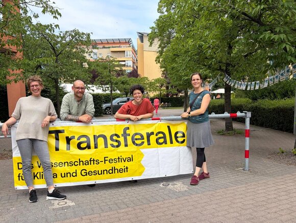 Ein gelbes Banner mit der Aufschrift "Transferale - Das Wissenschafts-Festival für Klima und Gesundheit". Daneben und dahinter stehen vier Personen: 3 Frauen und ein Mann
