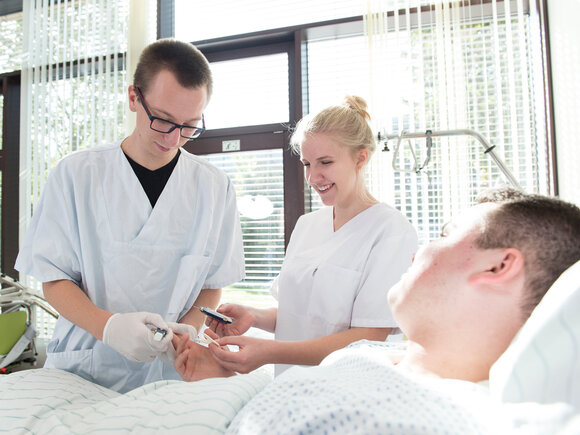 Ein junger Mann und eine junge Frau in weißen Kitteln versorgen eine pflegebedürftige Person im Bett.