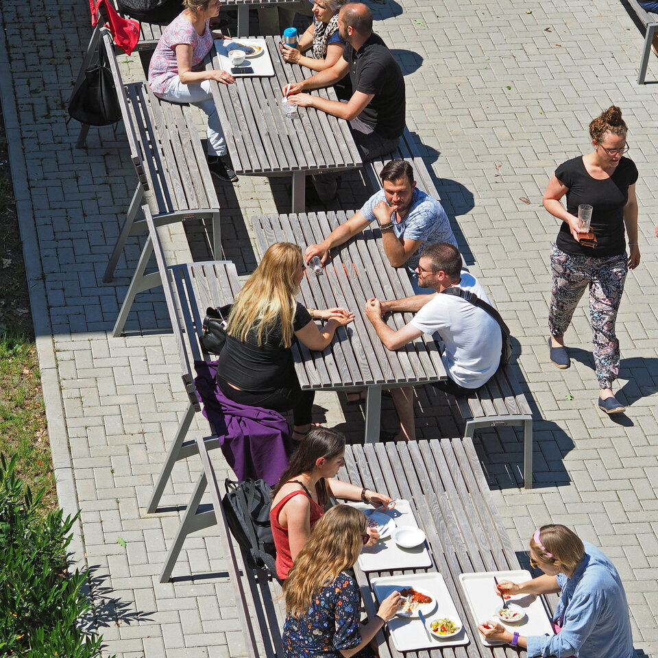 Blick auf die Außenbänke der Mensa, auf denen viele Studierende sitzen und sich unterhalten.
