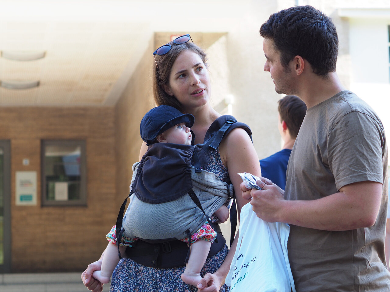 190614_KHSB_Vereinbarkeit_Heinrich_065 Eine Studentin mit Baby in der Trage unterhält sich mit einem Studenten vor dem Eingang der KHSB.