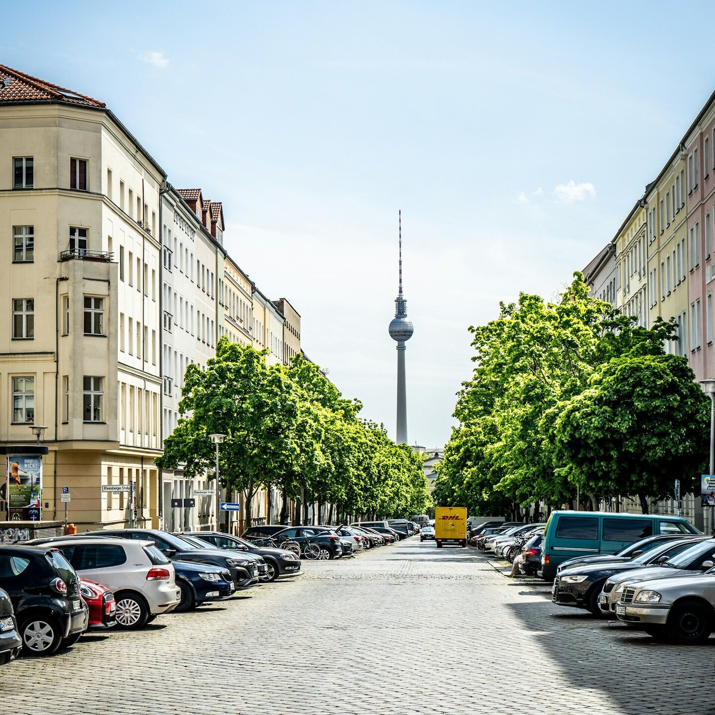 Der Berliner Fernsehturm steht am Ende einer Straßenflucht. Zu sehen sind leuchtend grüne Bäume, parkende Autos und Häuser