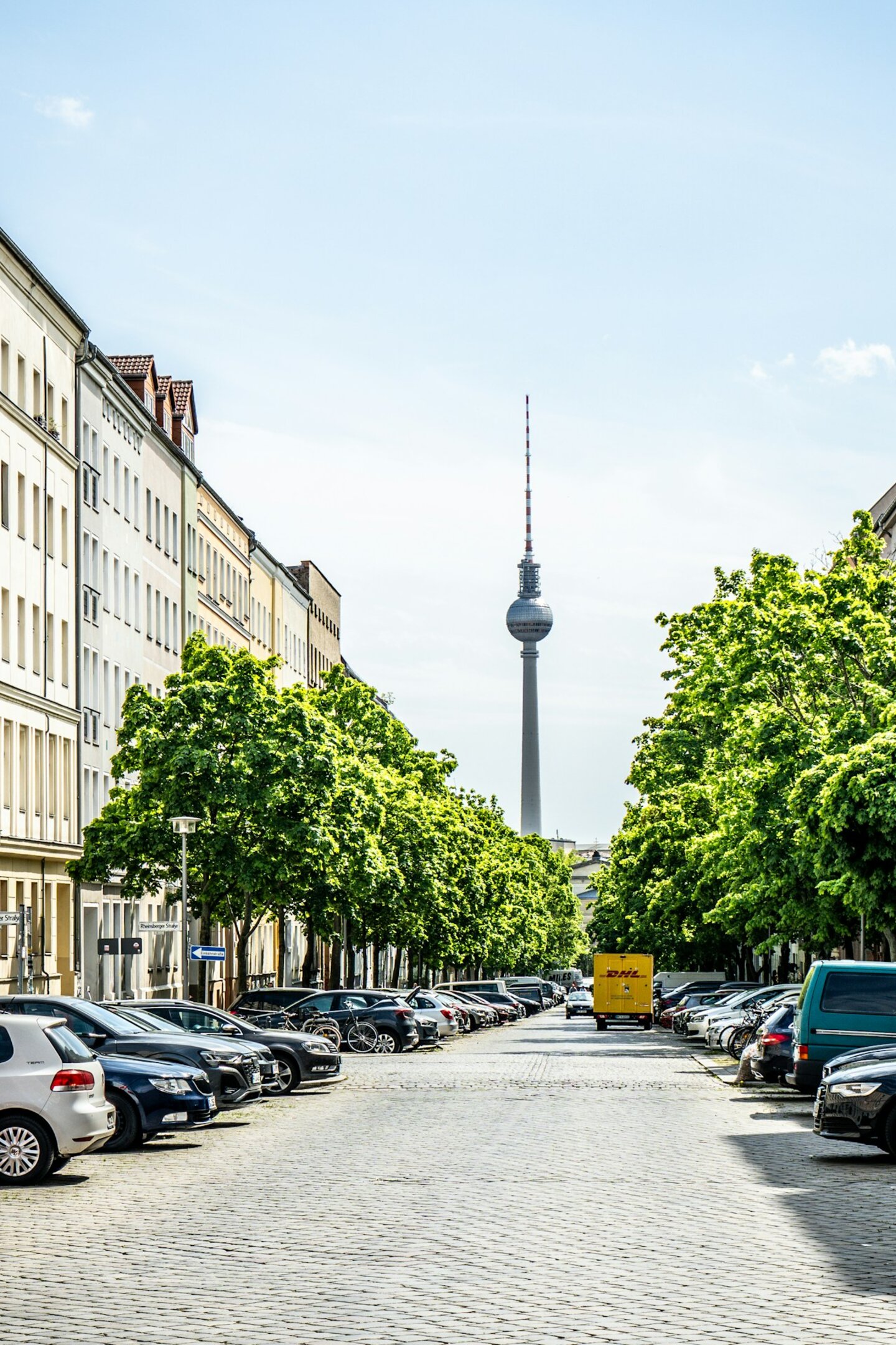 Der Berliner Fernsehturm steht am Ende einer Straßenflucht. Zu sehen sind leuchtend grüne Bäume, parkende Autos und Häuser