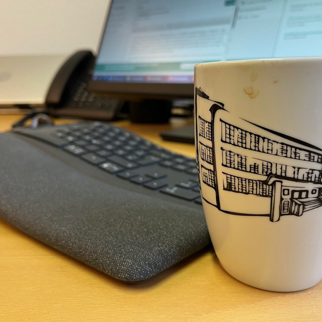 Foto mit Nahaufnahme eines Schreibtisches, im Vordergrund ist eine Tasse mit KHSB Werbeaufdruck, im Hintergrund sind Tastatur, Bildschirm und Laptop zu sehen.