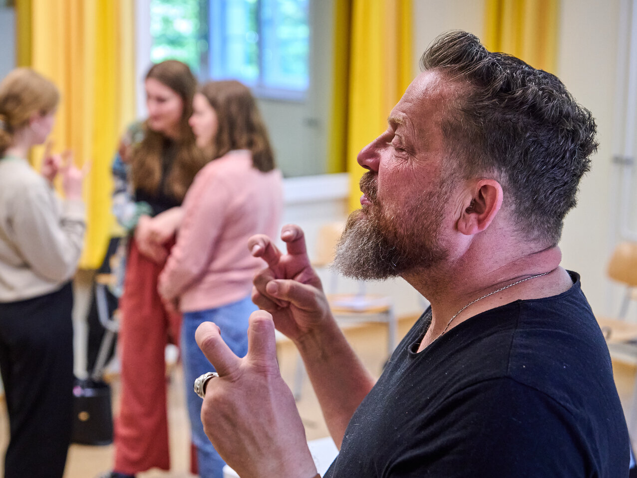Der Dozent ist im Vordergrund zu sehen, während eine dreiköpfige Gruppe von Studierenden im Hintergrund Gebärdensprache übt.
