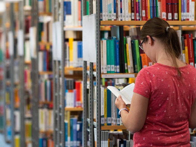 Eine Studentin steht zwischen den Regalen der Bibliothek und schaut in ein Buch.