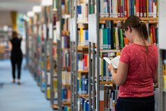 Eine Studentin steht zwischen den Regalen der Bibliothek und schaut in ein Buch.