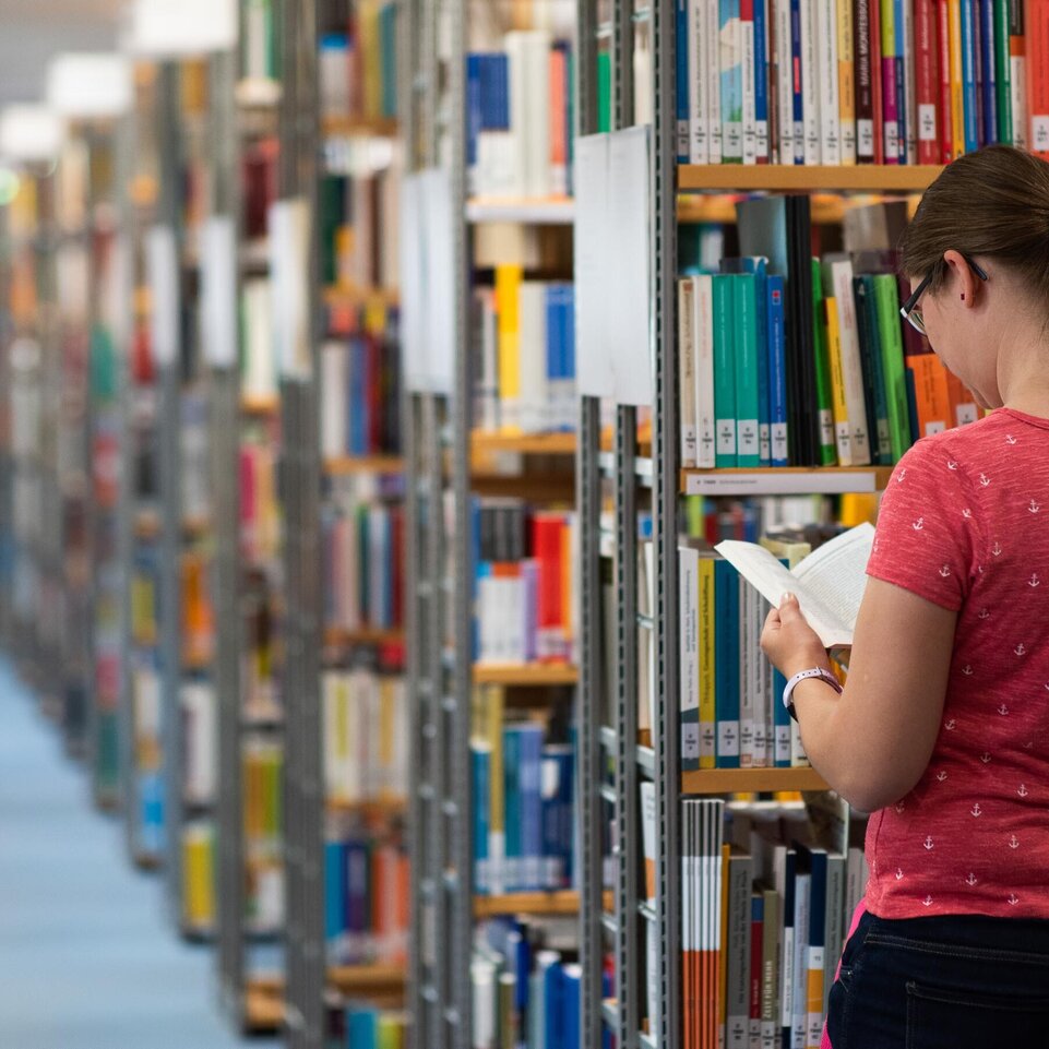 Eine Studentin steht zwischen den Regalen der Bibliothek und schaut in ein Buch.
