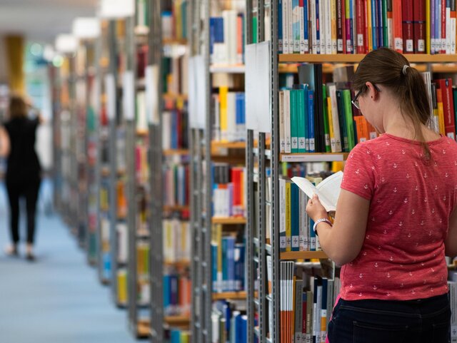 Eine Studentin steht zwischen den Regalen der Bibliothek und schaut in ein Buch.