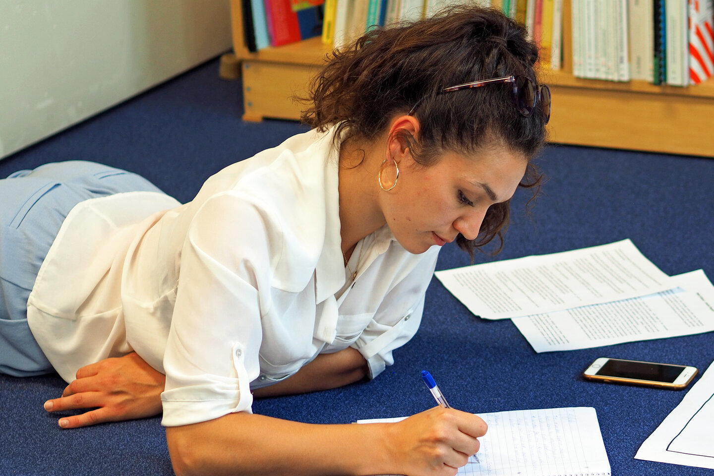 Eine Studierende liegt in der Bibliothek auf dem Boden und schreibt in ihr Notizbuch.