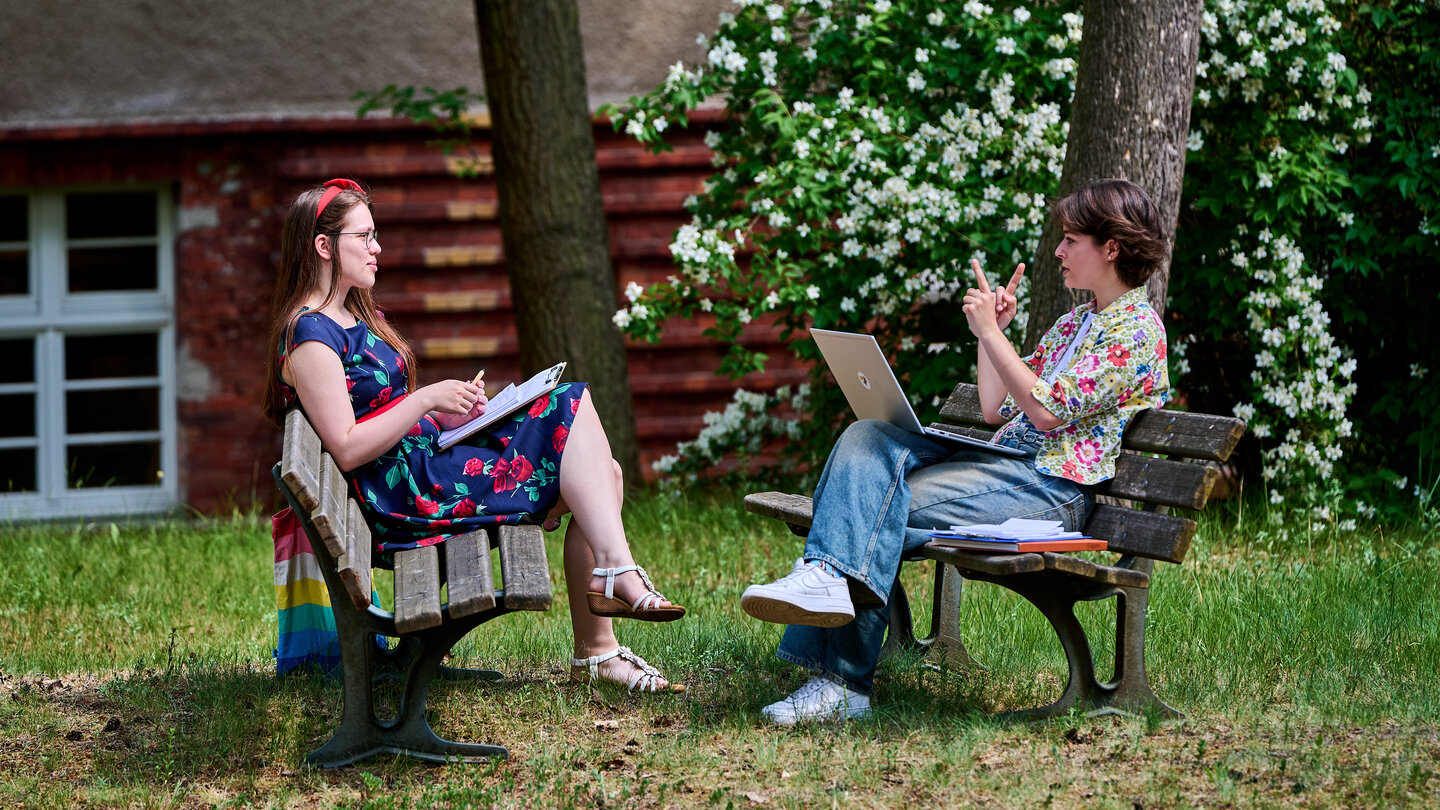 Zwei Studierende sitzen sich auf Bänken gegenüber und unterhalten sich.
