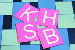 Auf Tafeln sind die vier Buchstaben K H S B dargestellt.