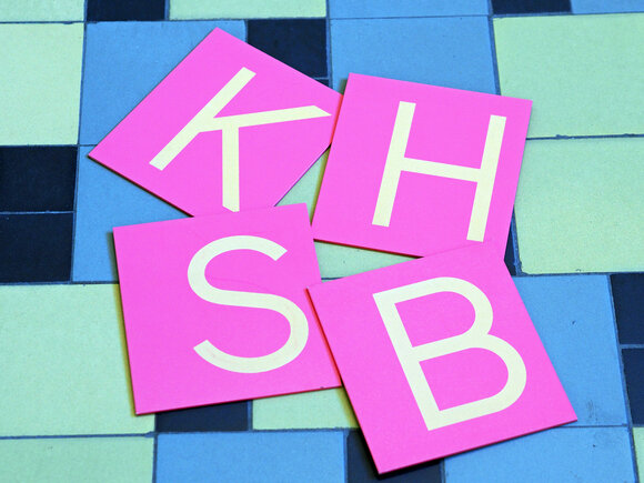 Auf Tafeln sind die vier Buchstaben K H S B dargestellt.
