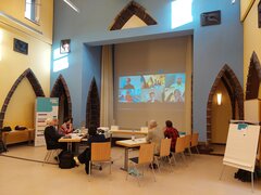 Zu sehen ist ein hoher Raum mit einer Leinwand mit Videokonferenz sowie sieben Personen an Tischen sitzend.