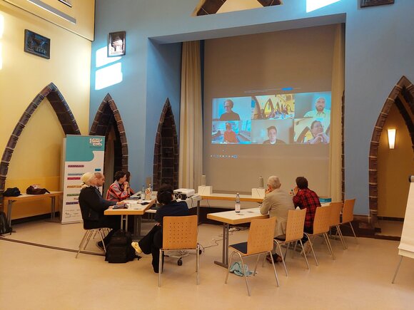 Zu sehen ist ein hoher Raum mit einer Leinwand mit Videokonferenz sowie sieben Personen an Tischen sitzend.