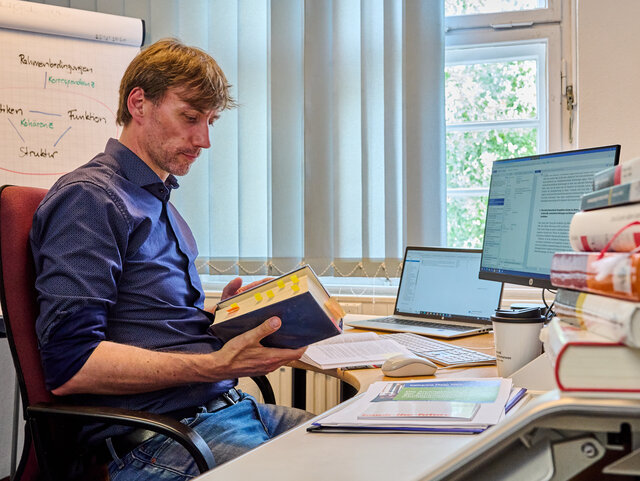 Ein Mitarbeiter der KHSB sitzt an seinem Schreibtisch und liest in einem Buch.
