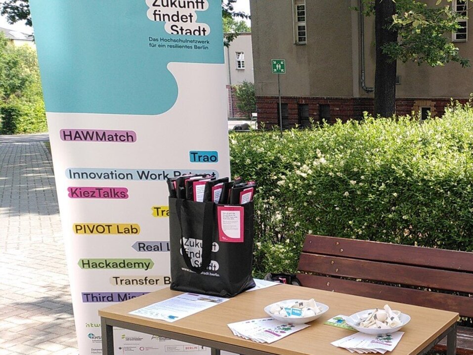 Hinter einem Tisch mit Infomaterialien steht ein Banner mit den Logos der einzelnen Teilvorhaben.