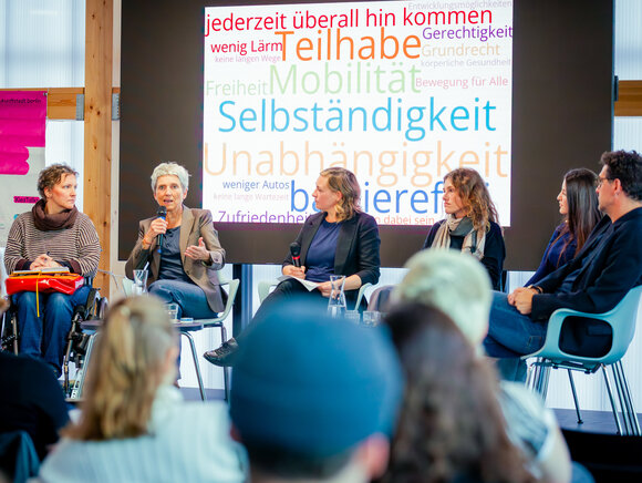 Zu sehen sind 6 Personen bei einer Diskussionsveranstaltung. Eine Person spricht in ein Mikrofon. Im Hintergrund sieht man eine Einblendung einer Wortwolke mit den Begriffen Selbstständigkeit, Teilhabe, Mobilität