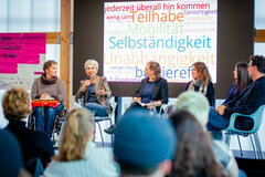 Zu sehen sind 6 Personen bei einer Diskussionsveranstaltung. Eine Person spricht in ein Mikrofon. Im Hintergrund sieht man eine Einblendung einer Wortwolke mit den Begriffen Selbstständigkeit, Teilhabe, Mobilität