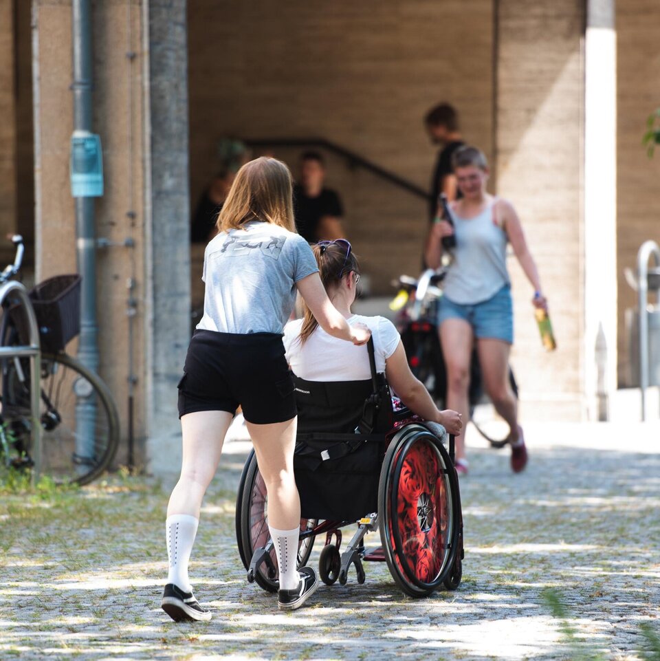 Eine Studentin begleitet eine Kommilitonin im Rollstuhl in Richtung Eingang der KHSB.