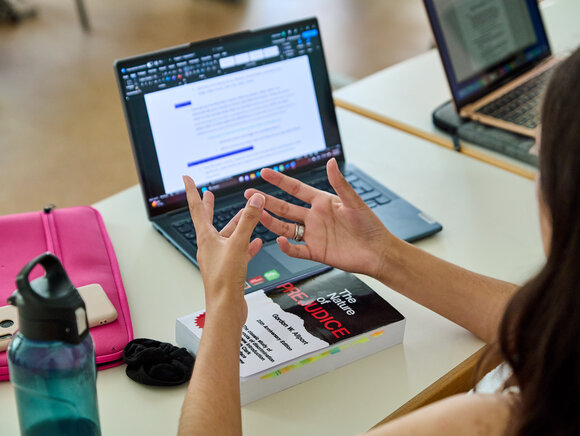 Ein Laptop steht auf dem Tisch, umgeben von Studienmaterialien, während eine Studierende gestikuliert.